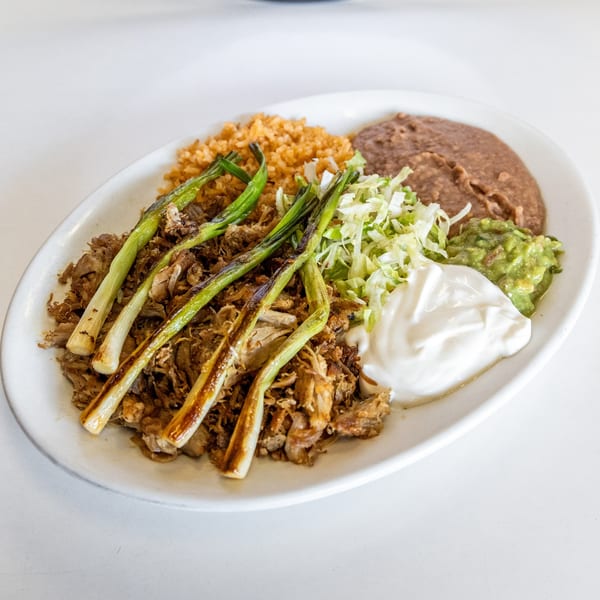 Mexican carnitas plate with grilled green onions, rice, refried beans, guacamole, and sour cream.