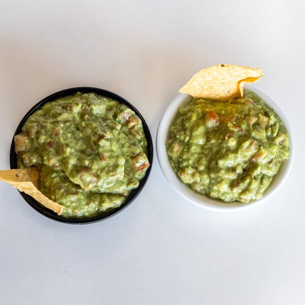 Two bowls of chunky guacamole with tortilla chips, one in a black bowl, one in a white bowl.